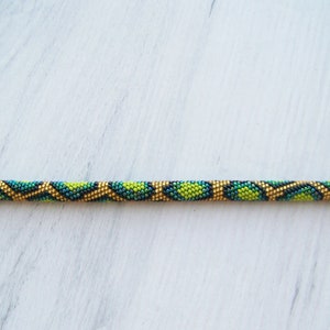 May include: A beaded pencil with a green, black, and gold geometric pattern. The pencil has metal end caps.