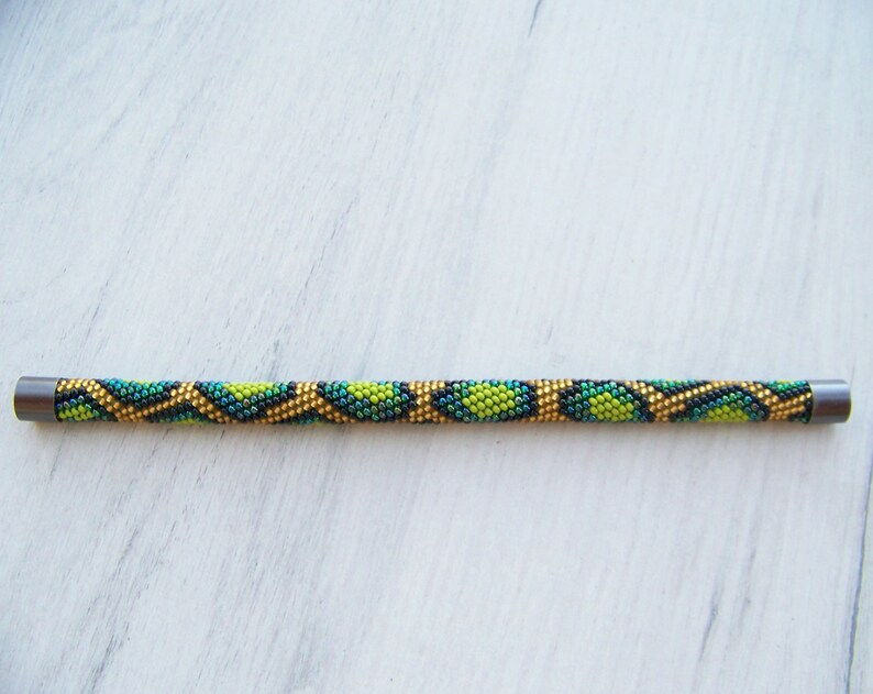 May include: A beaded pencil with a green, black, and gold geometric pattern. The pencil has metal end caps.