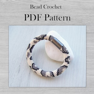 May include: A beaded crochet bracelet with a black, white, and gray snake skin pattern. The bracelet has a magnetic clasp and is resting on a white stone.