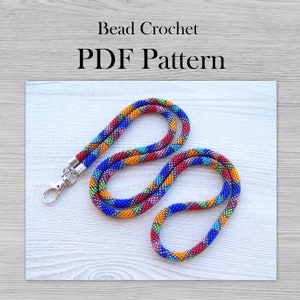 May include: A colorful beaded lanyard with a silver clasp. The lanyard is made with small beads in shades of blue, red, orange, green, and silver. The lanyard is arranged in a figure eight shape on a white wooden surface. The text "Bead Crochet PDF Pattern" is displayed above the lanyard.