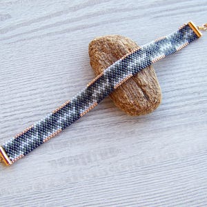 May include: A black, white, and gray beaded bracelet with a gold clasp and chain. The bracelet is on a wooden surface.