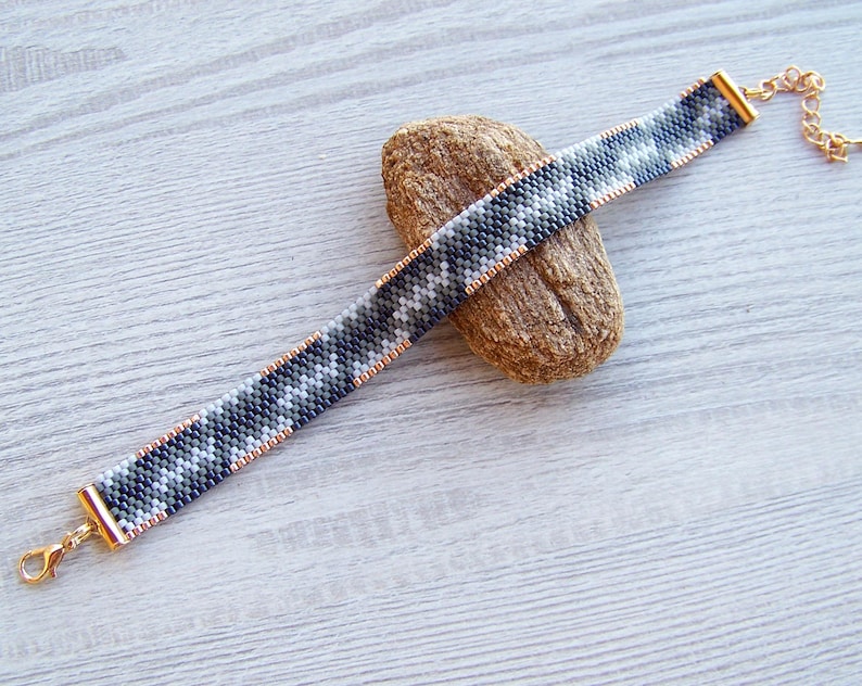 May include: A black, white, and gray beaded bracelet with a gold clasp and chain. The bracelet is on a wooden surface.