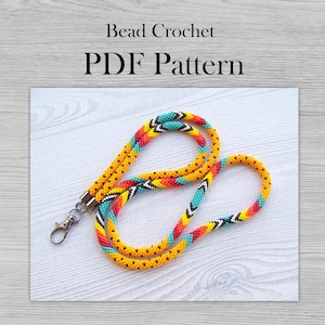 May include: A beaded lanyard with a black, white, yellow, orange, and turquoise geometric pattern. The lanyard has a silver clasp and is on a white wooden surface. Bead Crochet PDF Pattern.