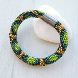 May include: A green, gold, and black beaded bracelet with a magnetic clasp. The bracelet is made of small, round beads that are arranged in a geometric pattern.