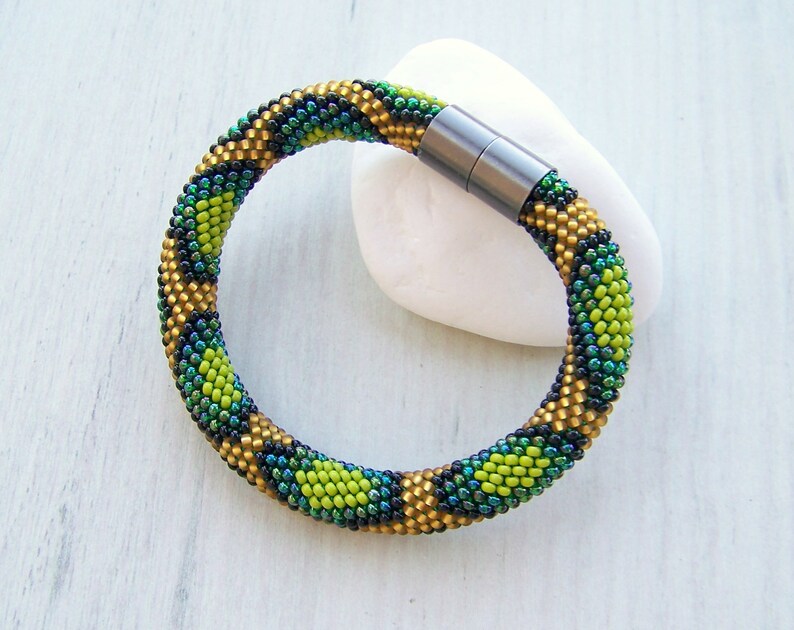 May include: A green, gold, and black beaded bracelet with a magnetic clasp. The bracelet is made of small, round beads that are arranged in a geometric pattern.