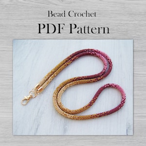 May include: A gold and burgundy beaded crochet necklace with a gold clasp. The necklace is on a white wooden surface. Bead Crochet PDF Pattern.