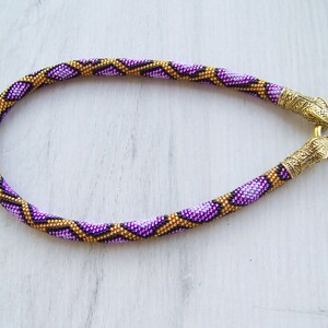 May include: A purple and gold beaded necklace with a snake-like pattern. The necklace has gold metal clasps on each end.
