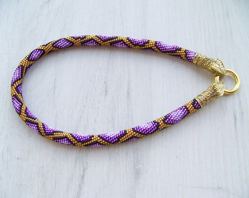 May include: A purple and gold beaded necklace with a snake-like pattern. The necklace has gold metal clasps on each end.