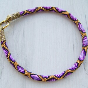 May include: A purple and gold beaded necklace with a decorative metal clasp. The necklace is made of small beads that are arranged in a geometric pattern.