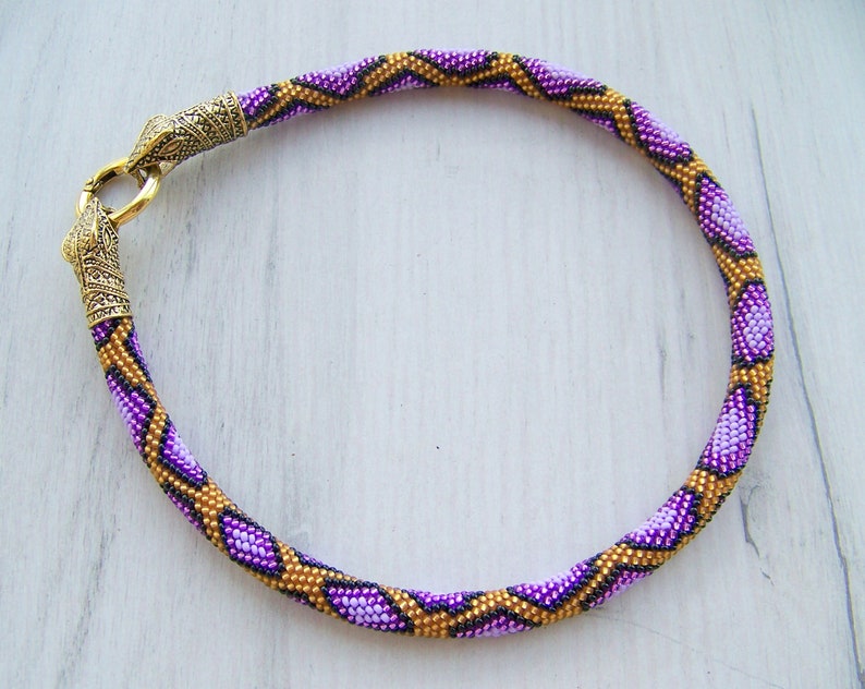May include: A purple and gold beaded necklace with a decorative metal clasp. The necklace is made of small beads that are arranged in a geometric pattern.