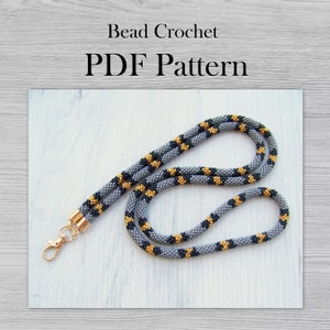 May include: A gray, black, and gold beaded necklace with a gold clasp. The necklace is on a gray wooden surface. Bead Crochet PDF Pattern.