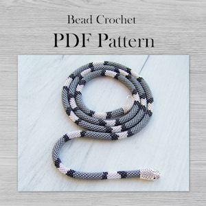 Peut inclure: Un collier de perles gris, noir et blanc avec un pendentif tête de serpent argenté. Le collier est enroulé sur une surface en bois blanc. Modèle de crochet de perles PDF.