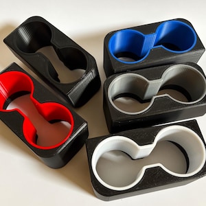 Puede incluir: Cinco portavasos impresos en 3D con anillos interiores de diferentes colores. Los portavasos son negros con anillos interiores rojos, azules, grises y blancos.