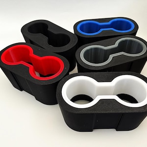May include: A selection of black car cup holders with coloured inserts. The inserts are red, blue, grey, and white. The cup holders are made of a textured material and are designed for a car's centre console.