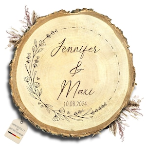 Op de afbeelding: Een ronde houten schijf met de namen "Jennifer & Maxi" en de datum "10.08.2024" gegraveerd. De namen staan in een bloemenkrans. Een label met "HERGESTELLT IN DEUTSCHLAND" is bevestigd.