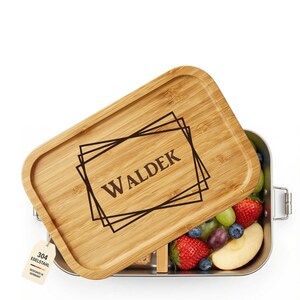 Könnte beinhalten: Eine Edelstahl-Lunchbox mit Bambusdeckel. Der Deckel ist mit dem Namen "WALDEK" in einem geometrischen Design graviert. Die Lunchbox ist mit frischem Obst gefüllt, darunter Erdbeeren, Trauben und Blaubeeren.