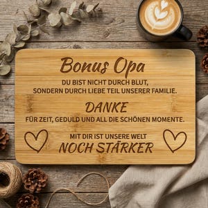Puede incluir: Una tabla de cortar rectangular de bambú con texto grabado a láser en alemán que dice: Bonus Opa, Du bist nicht durch Blut, sondern durch Liebe Teil unserer Familie. Danke für Zeit, Geduld und all die schönen Momente. Mit dir ist unsere Welt noch stärker, flanqueado por dos iconos de corazón.