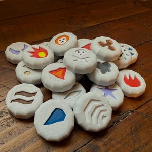 Pode incluir: Uma coleção de fichas de jogo redondas e brancas com vários símbolos e cores. Os símbolos incluem uma caveira e ossos cruzados, uma chama, uma estrela, uma onda e uma lua crescente. As fichas são feitas de um material texturizado e têm uma borda ligeiramente elevada.