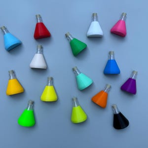 May include: A collection of colorful, miniature conical flasks with cork stoppers. The flasks are in various colors, including red, blue, green, yellow, and purple. The flasks are arranged on a light blue surface.