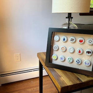 Op de afbeelding: Een zwarte, ingelijste schaduwdoos met 16 witte flessendoppen, elk met een ander geborduurd symbool in verschillende kleuren. De symbolen omvatten een golf, een regendruppel, een zon, een vuur, een ster, een wassende maan, een leeuw, een schedel, een hart, een vleermuis, een sleutel, een weegschaal, een wassende maan en een ster.