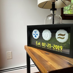 Op de afbeelding: Een zwarte, ingelijste schaduwdoos met drie witte ronde schijven met blauwe en gouden symbolen. De tekst "Est. 02-21-2019" wordt in geel op een zwarte achtergrond weergegeven.