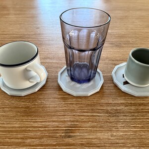 Op de afbeelding: Drie grijze onderzetters met een geometrisch ontwerp, elk met een andere beker. Een onderzetter houdt een witte keramische mok vast, een andere een helder glas en de derde een grijze keramische mok.