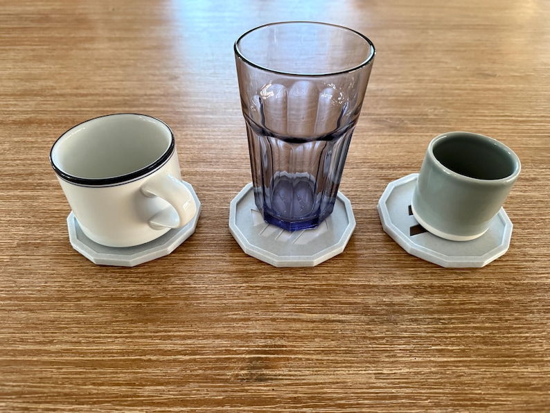 Op de afbeelding: Drie grijze onderzetters met een geometrisch ontwerp, elk met een andere beker. Een onderzetter houdt een witte keramische mok vast, een andere een helder glas en de derde een grijze keramische mok.