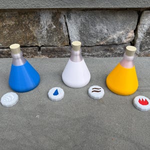 May include: Three colorful potion bottles with matching elemental symbols. The bottles are blue, white, and yellow, each with a light tan stopper. The symbols are water, earth, and fire.