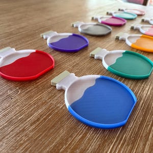 May include: A collection of colorful, potion-shaped coasters. Each coaster features a white and colored design, with a light green rectangular accent. Colors include red, purple, blue, green, orange, pink, gray, and teal. The coasters are arranged on a wooden surface.