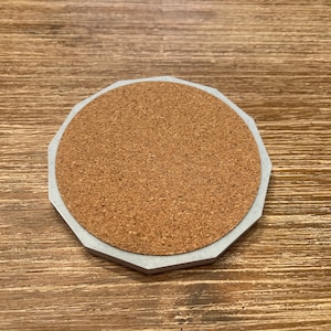 Op de afbeelding: Een ronde, grijze, betonnen onderzetter met een kurken inzet. De onderzetter heeft een licht verhoogde rand.