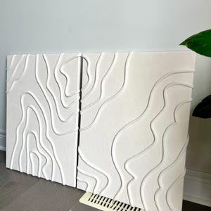 May include: Two white canvas paintings with a raised, textured design of white lines resembling topographic maps.