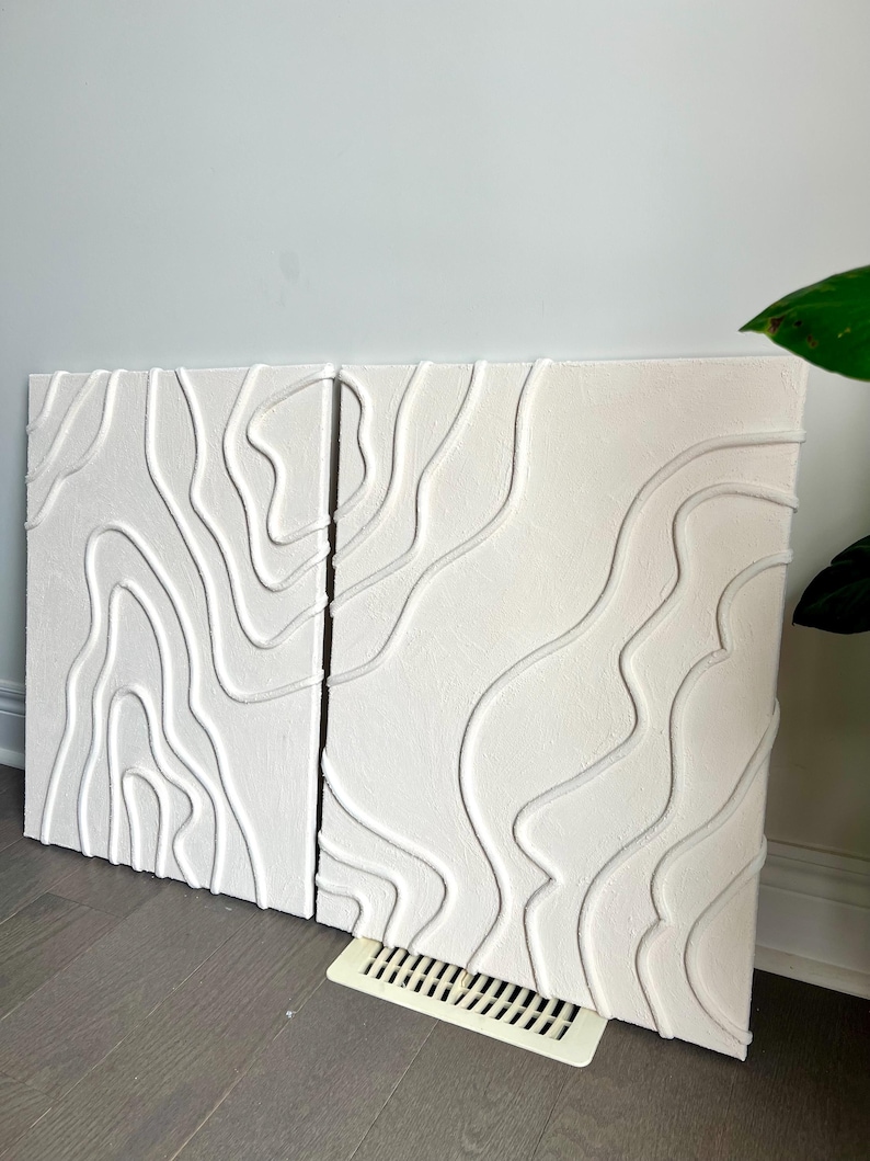 May include: Two white canvas paintings with a raised, textured design of white lines resembling topographic maps.