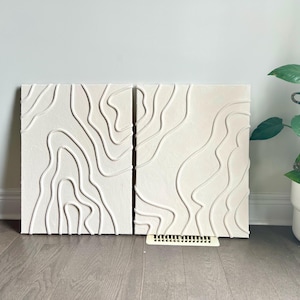 May include: Two white canvas wall art pieces with a raised, textured design of white lines resembling topographic maps.