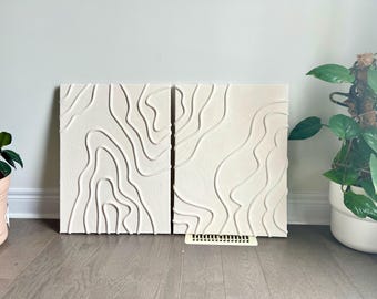 May include: Two white canvas wall art pieces with a raised, textured design of white lines resembling topographic maps.
