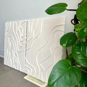 May include: Two white canvas panels with raised, textured lines in a wavy pattern. The panels are leaning against a wall, and a green plant is in the foreground.
