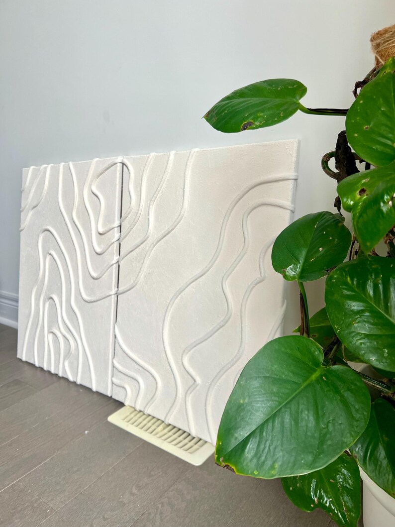May include: Two white canvas panels with raised, textured lines in a wavy pattern. The panels are leaning against a wall, and a green plant is in the foreground.