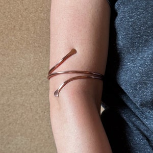 May include: A copper arm cuff with a simple design. The cuff is made of two thin pieces of copper wire that are twisted together. The ends of the wire are bent into small loops.