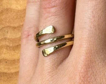May include: A gold-colored wrap ring with a hammered texture. The ring has a spiral design that wraps around the finger. The ring is shown on a hand with a wooden background.