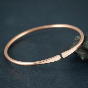 Op de afbeelding: Een eenvoudige koperen armband met een licht afgeronde vorm en een opening aan één kant voor gemakkelijk aan- en uittrekken.