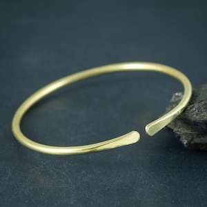 Può includere: Un bracciale aperto color oro. Il bracciale ha un design semplice e minimalista con una texture martellata. Le estremità aperte consentono una facile regolazione. Il bracciale è mostrato su uno sfondo scuro.
