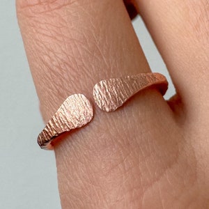 Può includere: Un anello regolabile in oro rosa con una superficie strutturata. L'anello ha un design aperto con due estremità curve a forma di foglia. L'anello è indossato su un dito.
