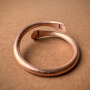 Op de afbeelding: Een koperkleurige, verstelbare ring met de letters "J" en "R" gegraveerd op de uiteinden. De ring heeft een glad, gebogen ontwerp en is geplaatst tegen een neutrale achtergrond.