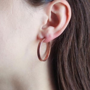 Könnte beinhalten: Nahaufnahme eines Ohrs einer Person, die ein Paar kupferfarbene Creolen trägt. Die Ohrringe sind kreisförmig und haben eine glatte, metallische Oberfläche. Die Person hat braunes Haar und helle Haut.