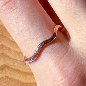 Op de afbeelding: Een gehammerde koperen ring met een golvend ontwerp. De ring wordt gedragen aan een vinger.