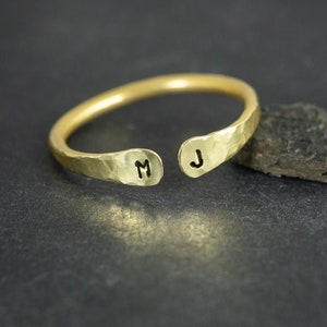 May include: A gold-coloured open ring with a hammered texture. The ring has the initials "M" and "J" stamped on the open ends. The ring is displayed on a dark surface next to a dark rock.
