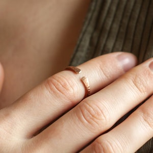 May include: A delicate, open-ended ring in a warm, rose gold hue. The ring features a textured band and is worn on a finger. The image is a close-up, showcasing the ring's design and the texture of the metal. The ring is a piece of jewelry.