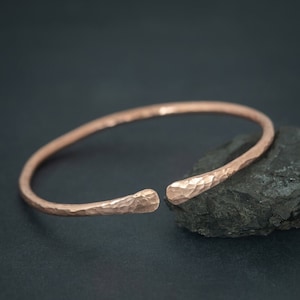 Puede incluir: Un brazalete de cobre martillado con un diseño ligeramente abierto. El brazalete es de estilo simple y minimalista y tiene un acabado metálico cálido.