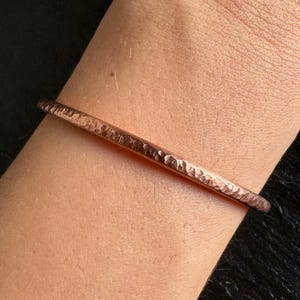 Può includere: Un braccialetto in rame martellato con una superficie strutturata. Il braccialetto ha un design semplice e minimalista e il rame ha una calda tonalità marrone-rossastra. Il braccialetto è mostrato sul polso di una persona.