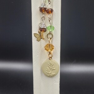 May include: A decorative bookmark with a gold-colored clasp and chain. The chain holds green, amber, and clear beads, a small gold butterfly charm, and a round, light green charm with a leaf design. The bookmark is white.