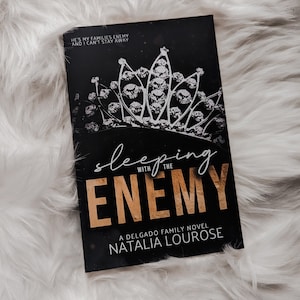 May include: A black book cover with a gold crown and the text "Sleeping with the Enemy" and "A Delgado Family Novel Natalia Lourose".